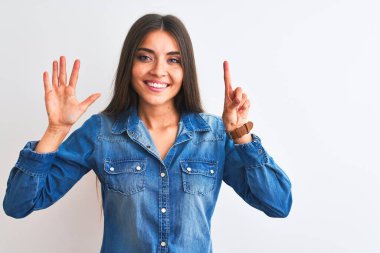 İzole edilmiş beyaz arka plan üzerinde duran sıradan bir kot gömlek giyen genç ve güzel bir kadın altı numaralı parmaklarıyla işaret ederken kendinden emin ve mutlu bir şekilde gülümsüyor..