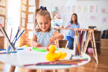 Beyaz kız çocuk, bayan öğretmenle oyun oynayıp öğreniyor. Anne ve kızı oyun odasında oyuncakların etrafında manyetik karatahtaya resim çiziyorlar.