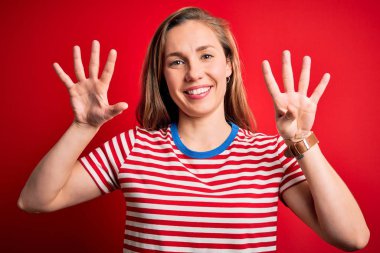 İzole edilmiş kırmızı arka plan üzerinde gündelik çizgili tişört giyen genç ve güzel sarışın kadın dokuz numarayı gösterirken kendinden emin ve mutlu bir şekilde gülümsüyor..