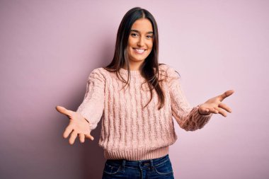 Young beautiful brunette woman wearing casual sweater over isolated pink background smiling cheerful offering hands giving assistance and acceptance.