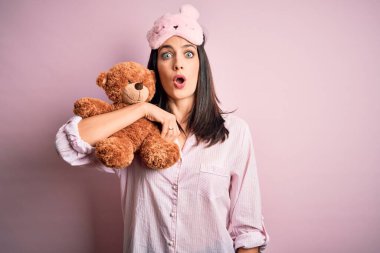 Young brunette woman with blue eyes wearing pajama hugging teddy bear stuffed animal scared in shock with a surprise face, afraid and excited with fear expression