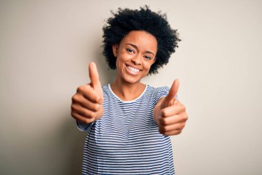 Young beautiful African American afro woman with curly hair wearing striped t-shirt approving doing positive gesture with hand, thumbs up smiling and happy for success. Winner gesture.