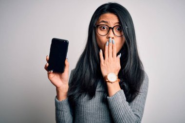 Young beautiful chinese woman holding smartphone over isolated white background cover mouth with hand shocked with shame for mistake, expression of fear, scared in silence, secret concept