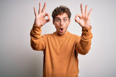 Sarışın, kıvırcık saçlı, beyaz arka planda sıradan bir süveter giyen, şaşırmış ve şok olmuş genç bir adam parmaklarıyla onay sembolü yapıyor. Çılgın ifade