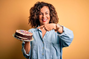 Middle age beautiful woman holding cake standing over isolated yellow background very happy pointing with hand and finger