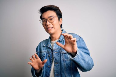 Beyaz arka planda kot ceket ve gözlük takan genç yakışıklı Çinli adam tiksindirici tepki verdiği için tiksindirici ve tiksindirici bir ifade takınıyor. Eller havada