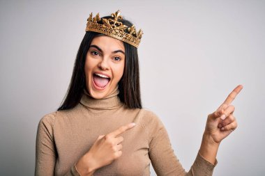İzole edilmiş beyaz arka planda altın kraliçe tacı takan genç ve güzel esmer kadın gülümsüyor ve kameraya bakıyor iki eliyle ve parmaklarıyla yan tarafı işaret ediyor..
