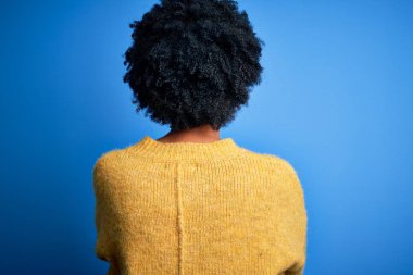 Young beautiful African American afro woman with curly hair wearing yellow casual sweater standing backwards looking away with crossed arms
