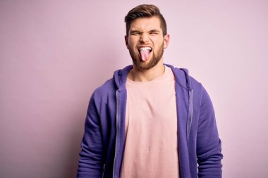 Sakallı, mavi gözlü, pembe arka planda mor eşofman üstlü, dilini mutlu bir ifadeyle dışarı çıkaran genç bir sarışın. Duygu kavramı.
