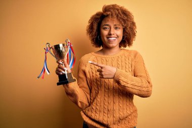 Kıvırcık saçlı, genç Afrikalı afro kadın şampiyonluk kupasını elinde ve parmaklarıyla mutlu bir şekilde tutuyor.