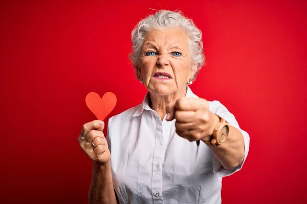 Senior beautiful woman holding paper heart standing over isolated red background annoyed and frustrated shouting with anger, crazy and yelling with raised hand, anger concept