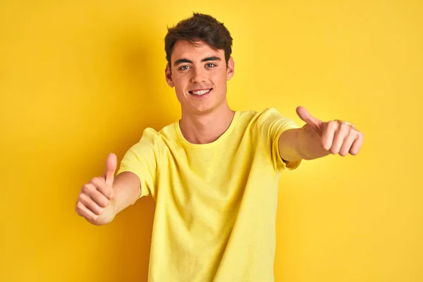 手で積極的なジェスチャーを行うことを承認孤立した背景に黄色のTシャツを身に着けている10代の少年 親指アップ笑顔と成功のために幸せ 勝者のジェスチャー — ストック写真