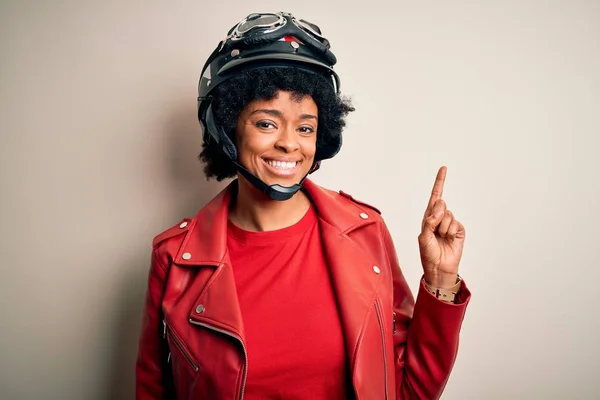 Young African American afro motorcyclist woman with curly hair wearing motorcycle helmet with a big smile on face, pointing with hand and finger to the side looking at the camera.