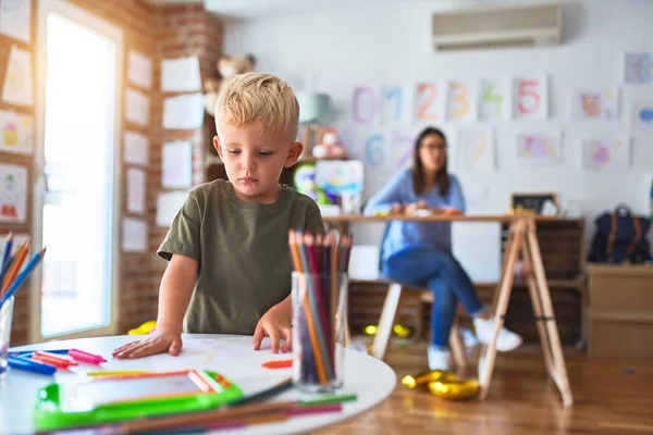 若い白人の子供は教師と一緒に学校で遊んでいる 母と息子は遊び場で色鉛筆で絵を描く若い女性は机の上に座っている — ストック写真