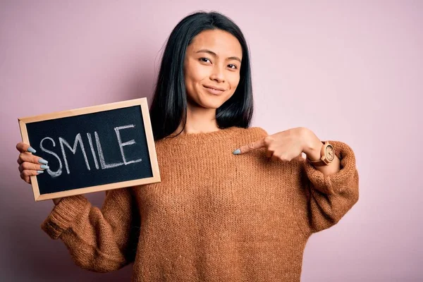 Young chinese woman holding blackboard with smile word over isolated pink background with surprise face pointing finger to himself
