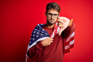 Gözlük takan genç adam ve Amerika Birleşik Devletleri bayrağı kızgın yüzüyle izole edilmiş kırmızı arka plan, olumsuz işaret baş parmakları aşağıda, reddedilme konsepti