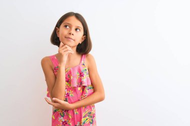 Genç, güzel, pembe çiçekli bir elbise giyen bir kız çocuğu. İzole edilmiş beyaz bir altyapının üzerinde duruyor. Elleri çenesinde, soru sormayı düşünüyor. Düşünceli bir yüzle gülümsüyor. Şüphe kavramı.