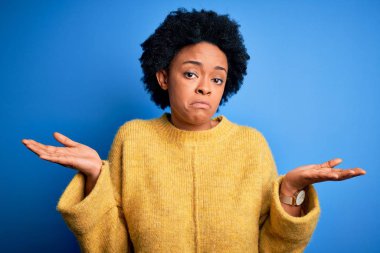 Young beautiful African American afro woman with curly hair wearing yellow casual sweater clueless and confused expression with arms and hands raised. Doubt concept.