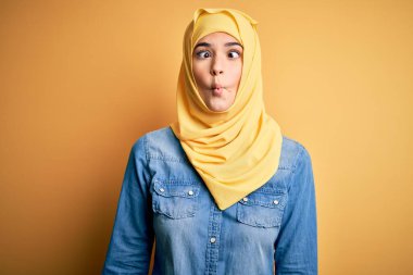 İzole edilmiş sarı arka planda duran Müslüman tesettürlü genç ve güzel bir kız dudaklı balık suratı yapıyor, çılgınca ve komik bir jest. Komik ifade.