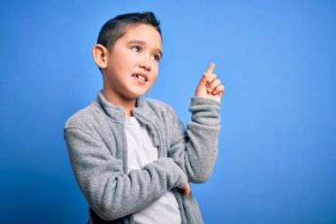 Küçük çocuk mavi izole edilmiş arka planda spor kazak giyiyor. Yüzünde kocaman bir gülümseme var. Eli ve parmağı kameraya bakıyor..