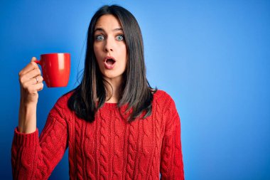 Young woman with blue eyes drinking cup of coffee standing over isolated background scared in shock with a surprise face, afraid and excited with fear expression