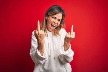 Orta yaşlı güzel sporcu kadın izole edilmiş kırmızı arka plan üzerine gündelik eşofman giyip orta parmak göstererek kötü ifade, provokasyon ve kaba tavır sergiliyor. Heyecanlı çığlık