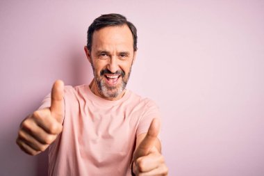 İzole edilmiş pembe arka planda duran günlük tişört giyen orta yaşlı beyaz bir adam elle olumlu jest yapmayı onaylıyor, gülümsüyor ve başarı için seviniyor. Kazanan jest.