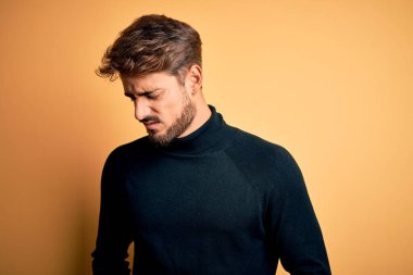 Balıkçı yaka kazak giyen sakallı genç yakışıklı adam midesi üzerinde sarı arka planda duruyor çünkü hazımsızlık, acı verici bir hastalık ve kendini iyi hissetmiyor. Ache kavramı.