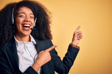 Afro saçlı, sarı arka planda kulaklık takan, gülümseyen ve kameraya bakıp iki eliyle ve parmaklarıyla yüzünü gösteren Afro Amerikalı genç bir kadın..