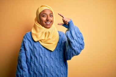 Genç Afrikalı Afro kadın, izole edilmiş sarı arka plan üzerine Müslüman başörtüsü takıyor gülümsüyor ve kendine güvenen el hareketleriyle küçük boy işareti yapıyor parmaklarıyla bakıyor ve kameraya bakıyor. Ölçü kavramı.