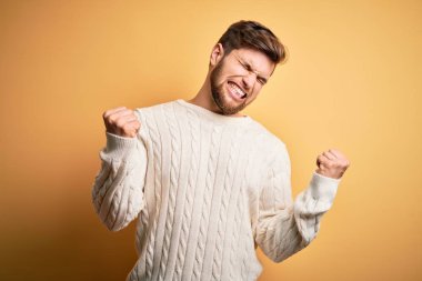 Sarışın, sakallı ve mavi gözlü genç adam sarı arka planda beyaz süveter giyiyor çok mutlu ve heyecanlı bir şekilde kollarını kaldırıp, gülümseyip başarı için çığlık atıyor. Kutlama konsepti.