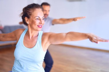 Orta yaşlı güzel sporcu çift mutlu gülümsüyor. Yüzünde gülümsemeyle ayakta durup, spor salonunda savaşçı pozu vererek yoga yapıyor.