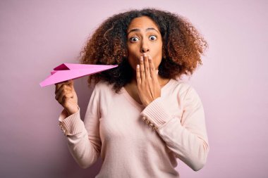 Afro saçlı, pembe arka planda kağıt uçakla gezen Afro-Amerikalı genç bir kadın. Ağzı kapalı, utancından ağzı açık, korku ifadesi, sessizlikten korkan, gizli bir konsept.