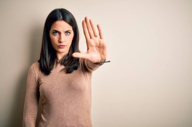 Young brunette woman with blue eyes wearing casual sweater over isolated white background doing stop sing with palm of the hand. Warning expression with negative and serious gesture on the face.