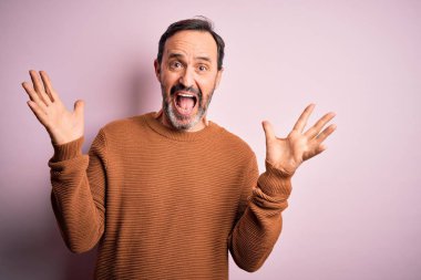 Orta yaşlı, beyaz, günlük kahverengi süveter giyen, izole edilmiş pembe arka plan üzerinde duran çılgın ve başarıyı kollarını kaldırıp heyecanla haykırarak kutlayan bir adam. Kazanan konsept
