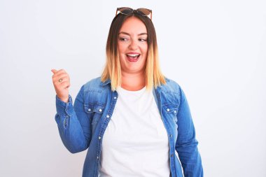 Güzel bir kadın kot gömlek giyiyor, izole edilmiş beyaz arka planda duruyor, gülümsüyor, gülümsüyor ve başparmağıyla yan tarafa bakıyor..
