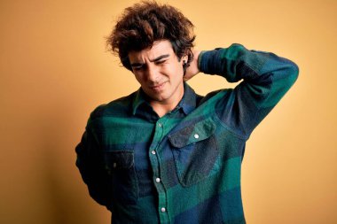 Genç, yakışıklı bir adam, günlük gömleğini giymiş, sarı arka planda dikiliyor boynu ağrıyor, boynuna elle dokunuyor, kas ağrısı çekiyor.