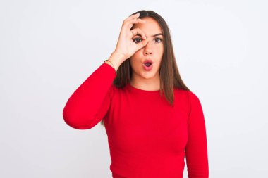 Young beautiful girl wearing red casual t-shirt standing over isolated white background doing ok gesture shocked with surprised face, eye looking through fingers. Unbelieving expression.