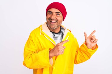 Yağmurluk ve yünlü şapka giyen orta yaşlı bir adam izole edilmiş beyaz arka planda duruyor gülümsüyor ve iki eliyle ve parmaklarıyla kameraya bakıyor..