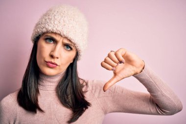 Young brunette woman with blue eyes wearing casual t-shirt and wool cap standing with angry face, negative sign showing dislike with thumbs down, rejection concept