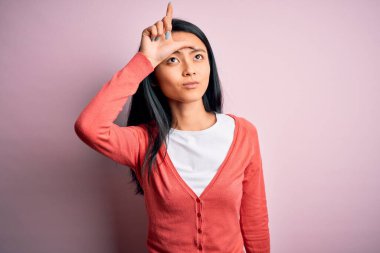 Young beautiful chinese woman wearing casual sweater over isolated pink background making fun of people with fingers on forehead doing loser gesture mocking and insulting.