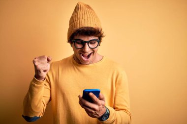 Genç yakışıklı adam, sarı arka planda gözlük takarak akıllı telefon kullanıyor. Gururla bağırıyor, zaferi ve başarıyı kutluyor. Çok heyecanlı, coşkulu bir duygu.