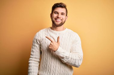 Sarışın, sakallı, mavi gözlü, sarı arka planda beyaz süveter giyen, yüzünde mutlu ve doğal bir ifadeyle el ve parmak gösteren bir gülümseme olan genç bir adam.