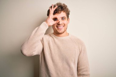 Sarışın, kıvırcık saçlı, beyaz arka planda sıradan bir süveter giyen, el hareketleriyle gülümseyen, mutlu yüzüyle parmaklarının arasından bakan genç, yakışıklı bir adam..