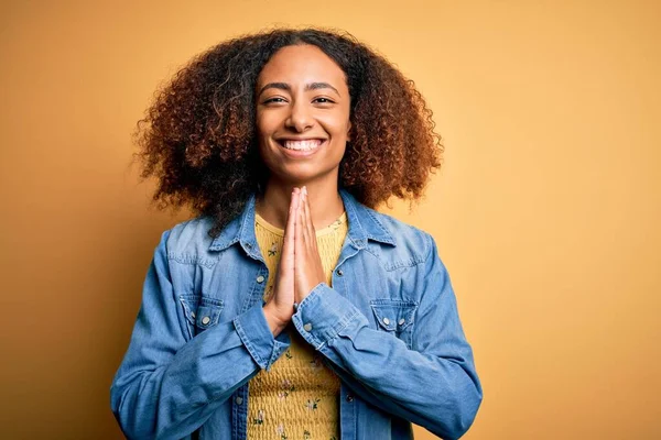 若いですアフリカ系アメリカ人女性とともに手で祈って黄色の背景にカジュアルなデニムシャツを身に着けているアフロの髪とともに許しを求めて笑顔自信 — ストック写真