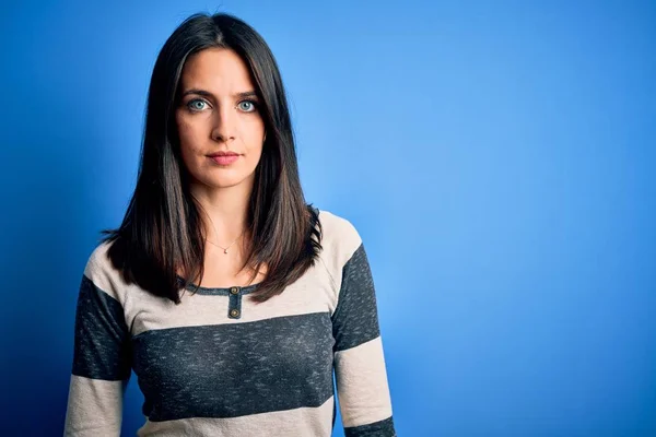 Young brunette woman with blue eyes wearing casual striped t-shirt standing over background with serious expression on face. Simple and natural looking at the camera.