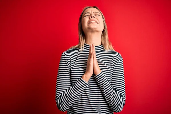 Young beautiful blonde woman wearing casual striped sweater over red isolated background begging and praying with hands together with hope expression on face very emotional and worried. Begging.