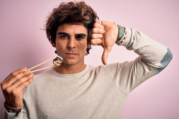若いハンサムな男は怒っている顔で孤立したピンクの背景の上に箸を使用して寿司を食べて 否定的なサインは親指を下げて嫌いを示す 拒否の概念 — ストック写真