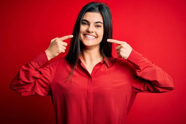 若い美しいブルネットの女性は 赤い背景の上に立ってカジュアルなシャツを着て陽気に見せ 指の歯と口で指して笑っています 歯科衛生の概念 — ストック写真