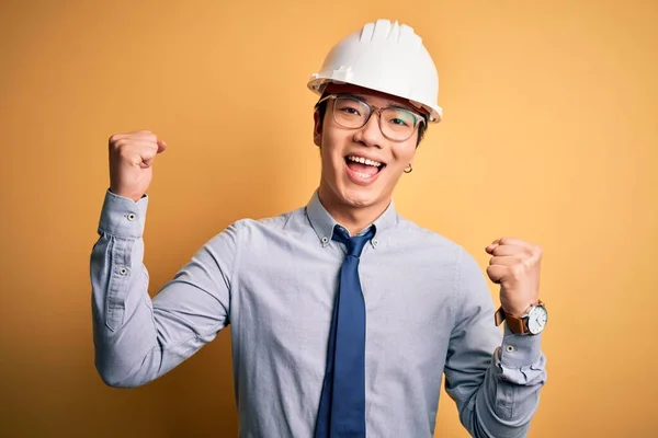 年轻英俊的中国建筑师 戴着安全帽 系在黄色的背景上 以张开双臂和睁开眼睛庆祝成功 赢的概念 — 图库照片